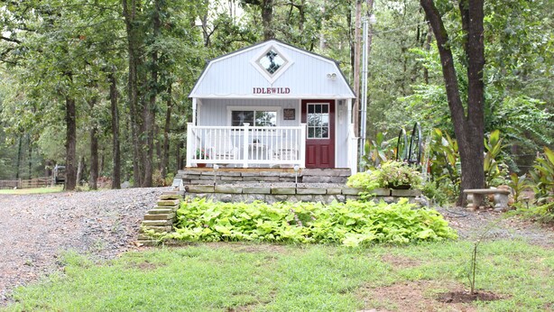A small, white building with a red door and a balcony, surrounded by lush greenery and trees. - Little House in the Pine Woods (Heavener)