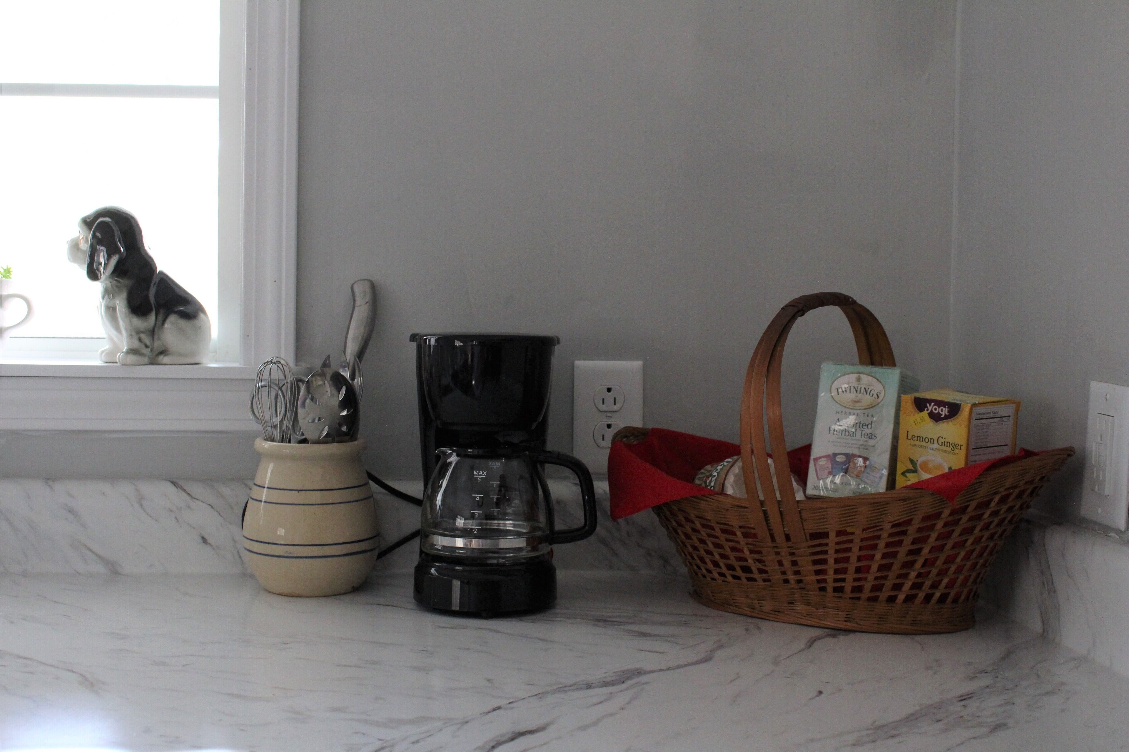 A kitchen counter with a coffee maker, a basket of groceries, and a ceramic pot with utensils.