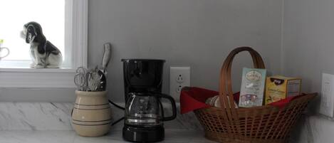 A kitchen counter with a coffee maker, a basket of groceries, and a ceramic pot with utensils.