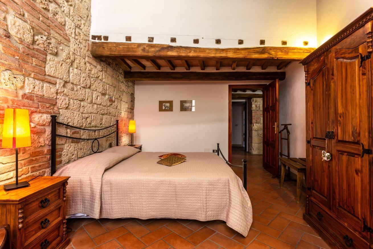 A bedroom with a brick wall, wooden ceiling beams, a bed with a beige quilt, a nightstand with a lamp, and a wooden wardrobe.