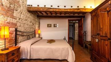 A bedroom with a brick wall, wooden ceiling beams, a bed with a beige quilt, a nightstand with a lamp, and a wooden wardrobe.