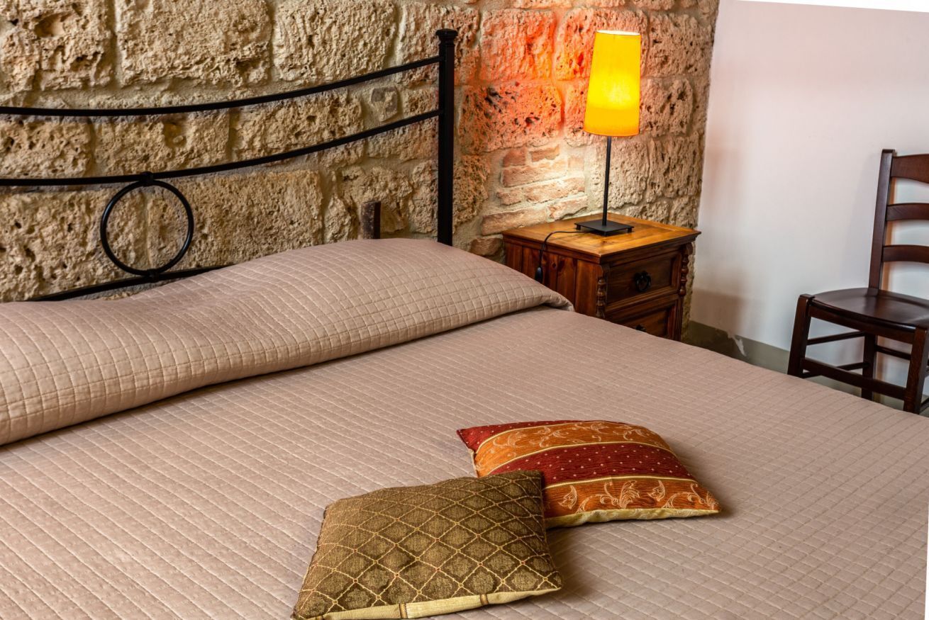 Una cama con cubierta acolchada, dos almohadas, una mesita de noche con lámpara y una silla de madera.