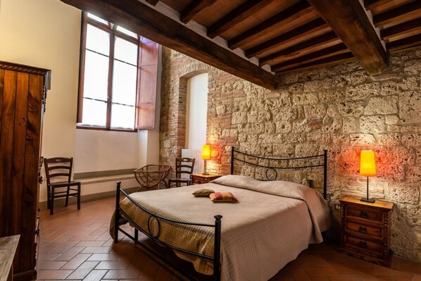 Una camera da letto rustica con parete in pietra, soffitto in legno, un letto con struttura in metallo, due comodini con lampade e una finestra con persiane.