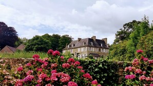 En stor bygning af sten med flere skorstene, omgivet af frodig vegetation og blomstrende planter.
