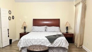 Un dormitorio con una cama grande, dos mesitas de noche con lámparas, un otoman redondo y una puerta.