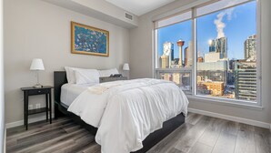 Une chambre moderne avec un grand lit, deux tables de chevet, une commode, une œuvre d’art encadrée et une vue sur la ville.