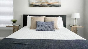 Un lit bien fait, recouvert d’une couette blanche, avec une tête de lit sombre et un oreiller à motifs bleus. Au-dessus du lit, on aperçoit un tableau, et sur la table de chevet, une petite plante et une lampe.