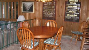 Een houten eethoek met een ronde tafel en zes stoelen, een hanglamp, een lijst met het menu aan de muur en een gestreepte bank op de achtergrond.