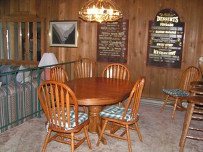A wooden dining area with a round table and six chairs, a hanging light fixture, a framed menu on the wall, and a striped sofa in the background.