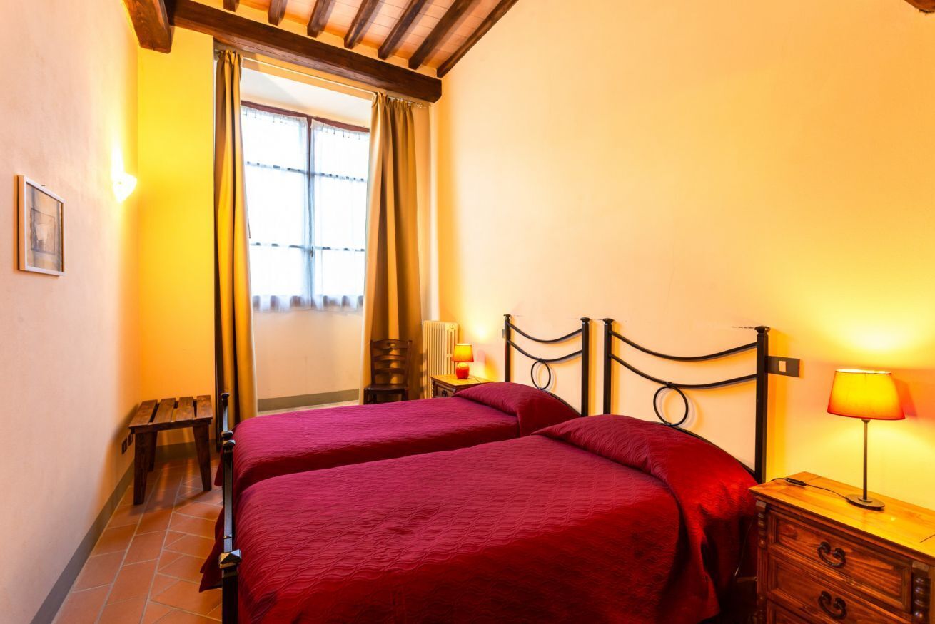 Habitación con dos camas, cabeceros de madera, un banco, una mesita de noche con lámpara y una ventana con cortinas.