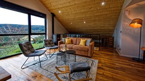 A spacious loft with a high ceiling, large windows offering a view of the countryside, a cozy seating area with a sofa and chairs, and a dining table.