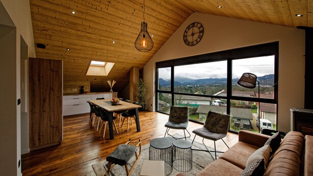 A spacious living area with a wooden ceiling, a large window offering a view of the outdoors, a cozy seating area with a sofa and chairs, and a dining space with a table and chairs.