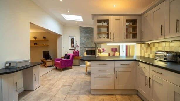 Una cocina moderna con armarios de madera clara, encimera negra y un tostador empotrado. Hay una zona de estar con una silla rosa y una mesita.