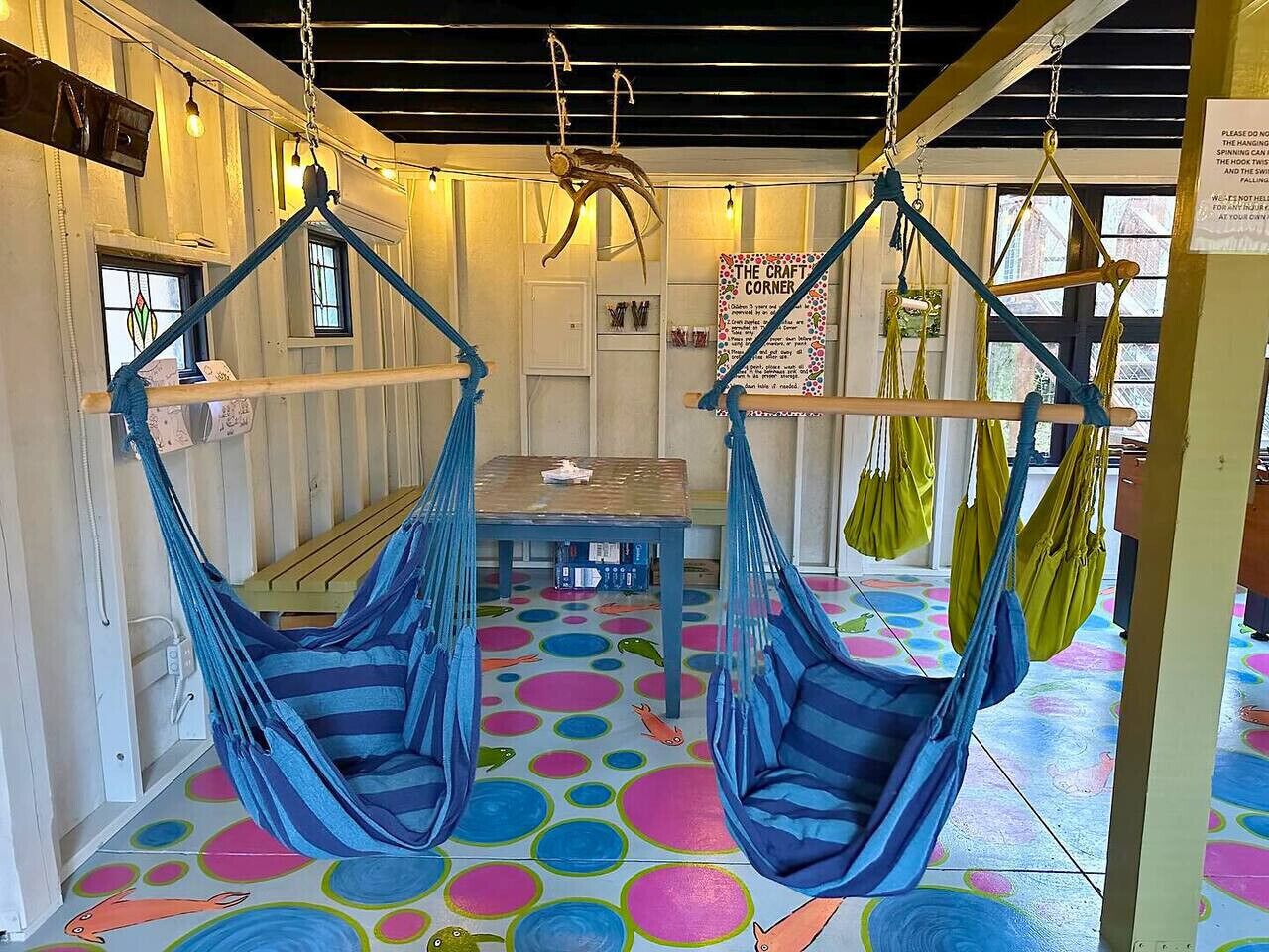 A room with two blue hammocks suspended from the ceiling, a wooden table in the center, and a colorful circular floor pattern.