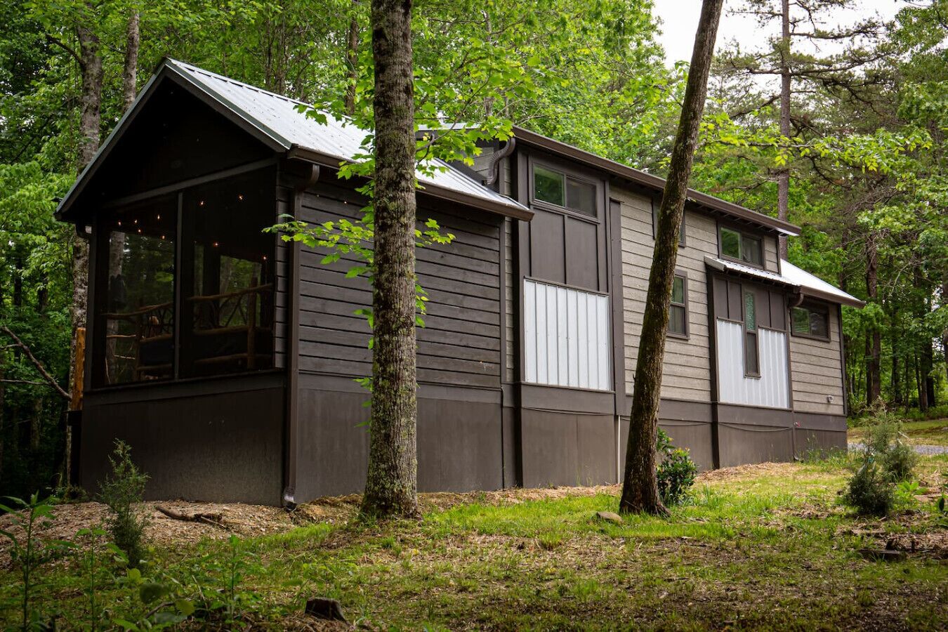 Une cabane dotée d’un toit à pignon, de grandes fenêtres et d’une façade en bois, entourée d’arbres.