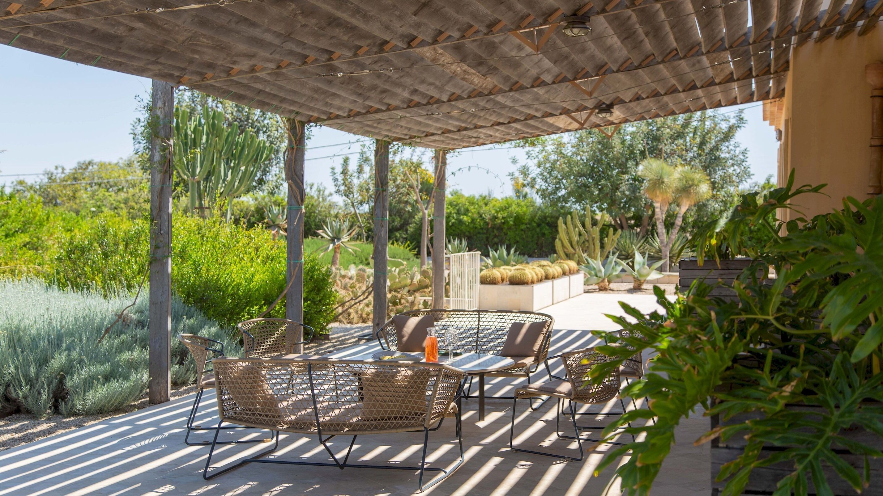 An outdoor patio with wicker furniture, a pergola, and a variety of plants.