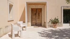 A building entrance with a wooden double door, a potted cactus, and two white chairs outside. - Villa Majoli 9, Emma Villas (Noto)