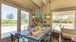 A dining area with a large table, chairs, hanging blue pendant lights, and a bookshelf filled with books. - Villa Majoli 9, Emma Villas (Noto)