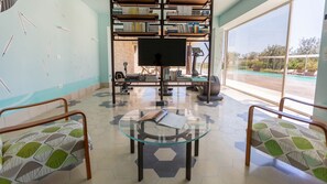 A reading area with a glass table, two chairs, a bookshelf, and a large window with a view of a pool. - Villa Majoli 9, Emma Villas (Noto)