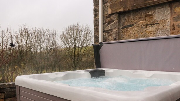 A hot tub with a stone wall backdrop and a view of leafless trees. - Coach House Cottage (Carnforth)