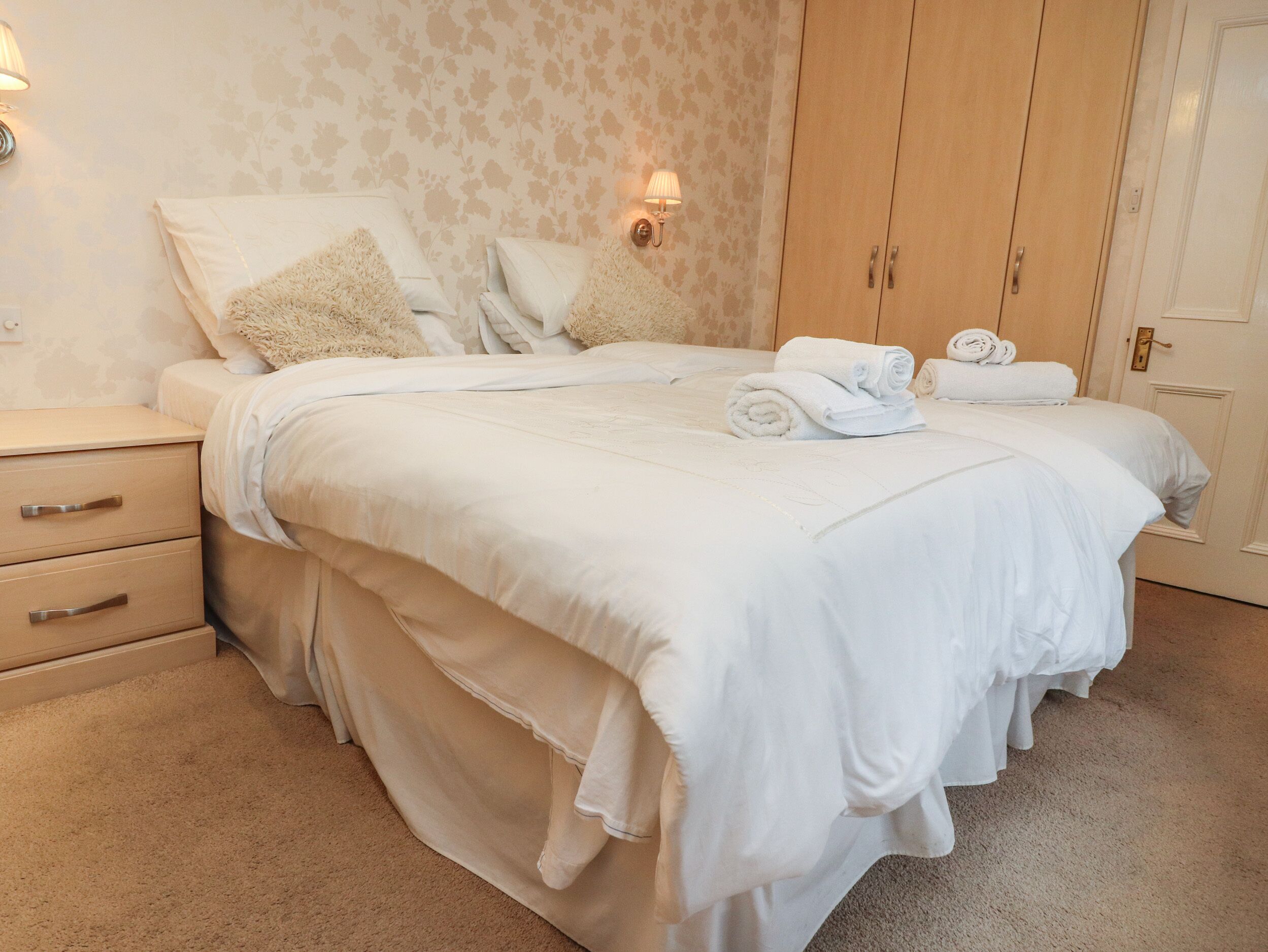 A neatly made bed with white linens and pillows, a wooden nightstand with drawers, and a door with a light switch.