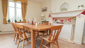 A dining room with a wooden table and chairs, a fireplace with a mantel decorated with festive garlands, and a window with curtains. - Coach House Cottage (Carnforth)