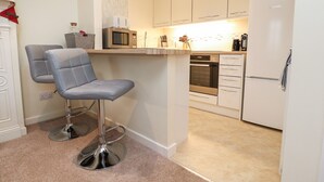 A modern kitchen with white cabinets, a microwave, an oven, and a refrigerator. There are two bar stools with metal bases. - Coach House Cottage (Carnforth)