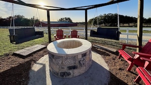A covered outdoor seating area with swings and Adirondack chairs, a central fire pit, and a view of a fenced field.