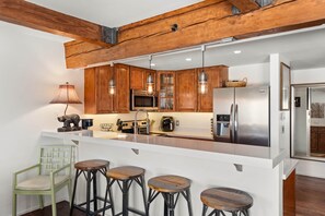 Una cocina moderna con armarios de madera, un refrigerador de acero inoxidable, un microondas y una cafetera. Hay tres taburetes y una silla.