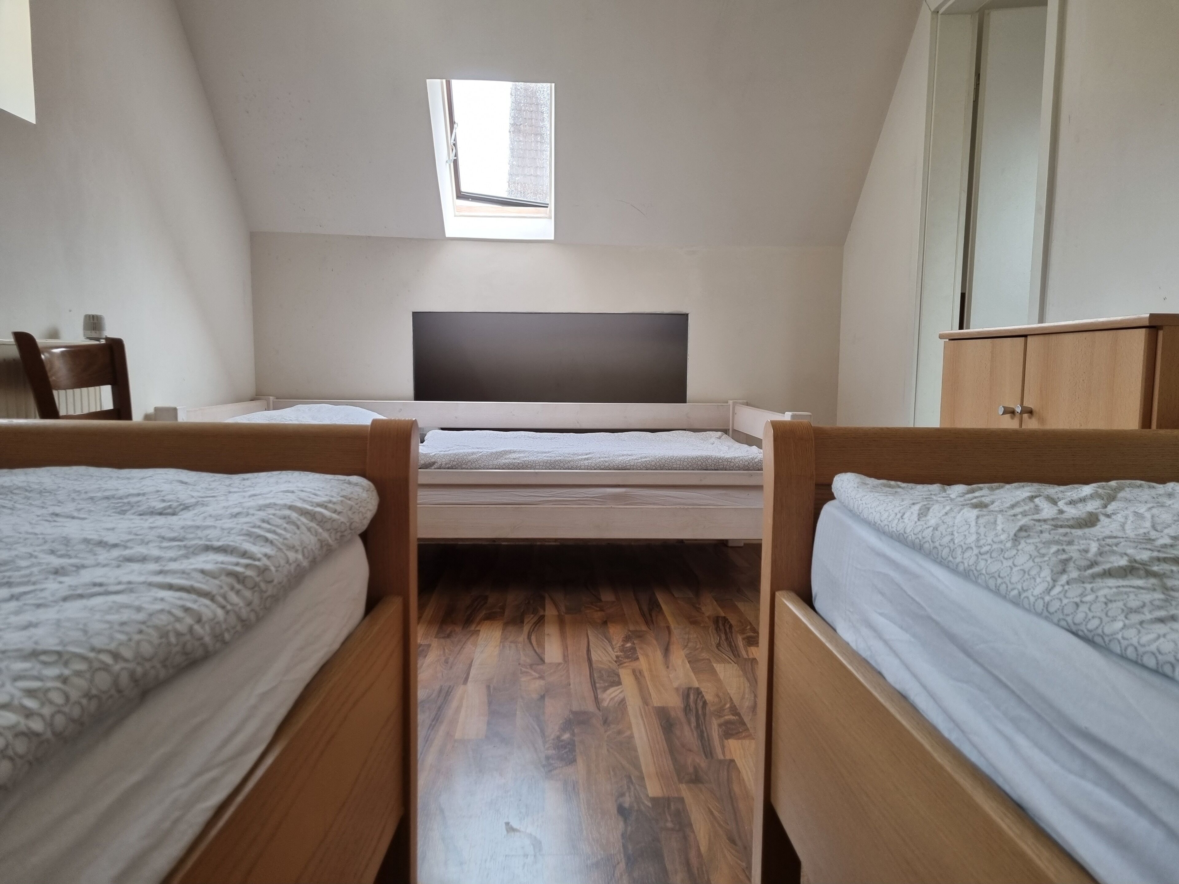 A dormitory room with two wooden bunk beds, a small window, a desk, and a wardrobe.