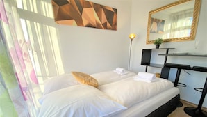 Moderni hotellihuone, jossa on suuri sänky, yöpöytä lampun kanssa, peili ja geometrisesti kuvioitu seinätaideteos.