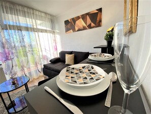 Een moderne woonkamer met een eethoek. Er is een zwarte bank, een glazen salontafel en een eettafel met borden en bestek. Een schilderij hangt aan de muur en er is een raam met gordijnen.