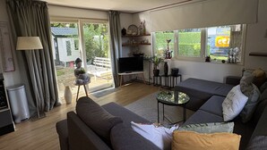 A modern living room with a grey sofa, a small coffee table, and a TV. There is a window with curtains, a bookshelf, and a small table with decorative items.