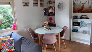 A cozy living room with a grey sofa, a round dining table with pink chairs, a white wall clock, and a shelf with various items.
