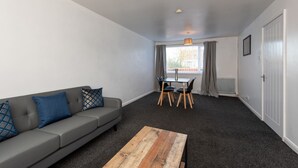Ein modernes Wohnzimmer mit grauem Sofa, einem Holztisch, einem Esstisch mit Stühlen und einem Fenster mit Vorhängen.