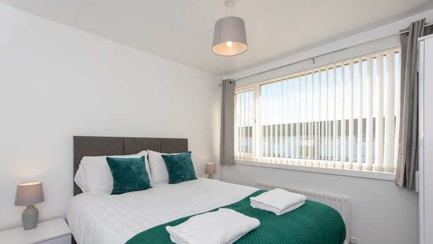 Ein Hotelzimmer mit einem Bett, einem Nachttisch, einer Lampe, einem Fenster mit Jalousien und Blick ins Freie.