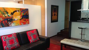 Un salon moderne avec un canapé en cuir noir, des coussins rouges et noirs, une table basse en verre et un tapis rouge. Un tableau est accroché au mur, et on aperçoit une cuisine au fond.