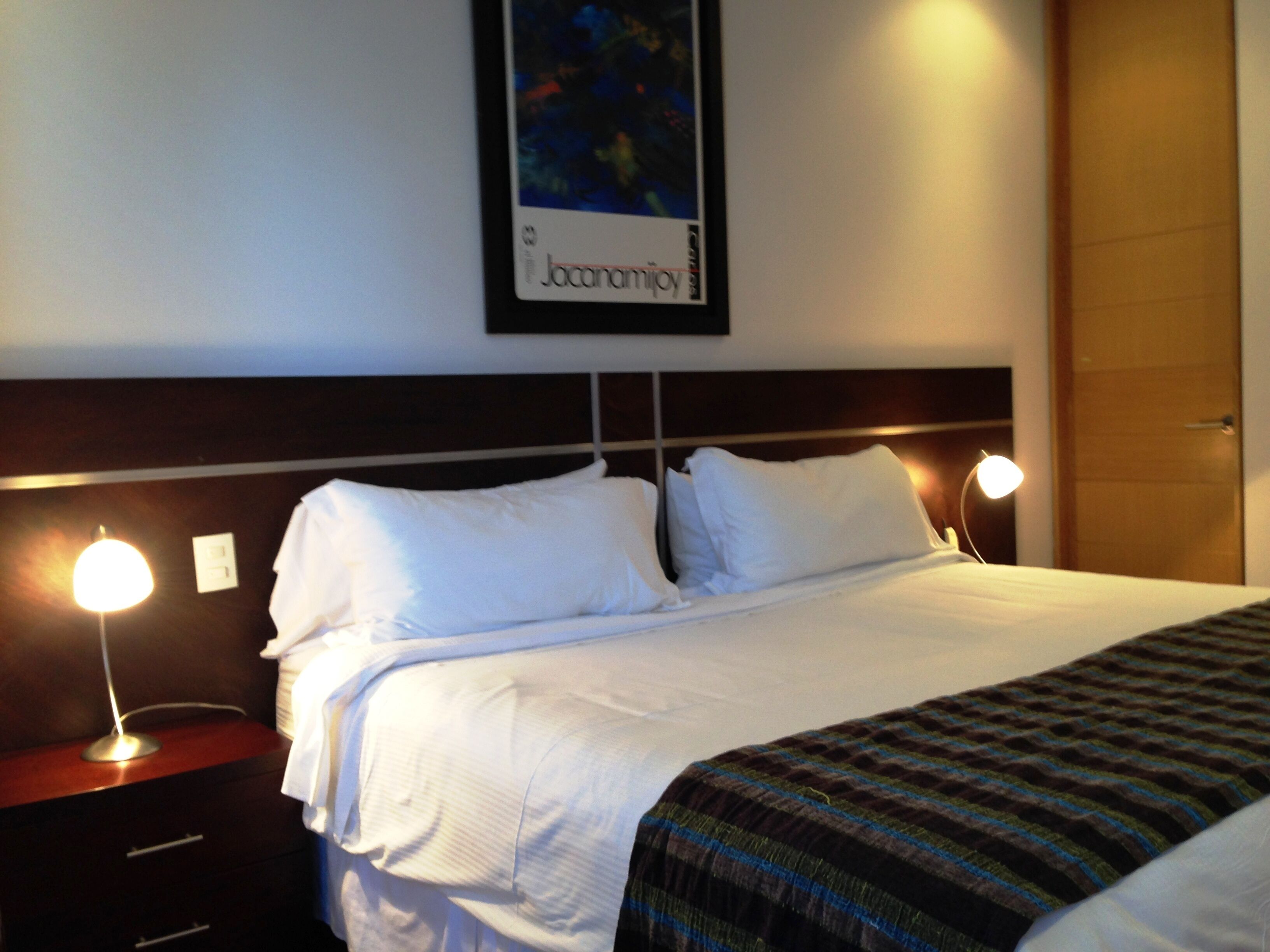 A hotel room with a neatly made bed, two bedside lamps, a nightstand, and a framed picture on the wall.