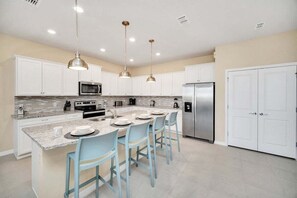 Una cocina moderna con armarios blancos, una isla central con taburetes de bar, electrodomésticos de acero inoxidable e iluminación colgante.