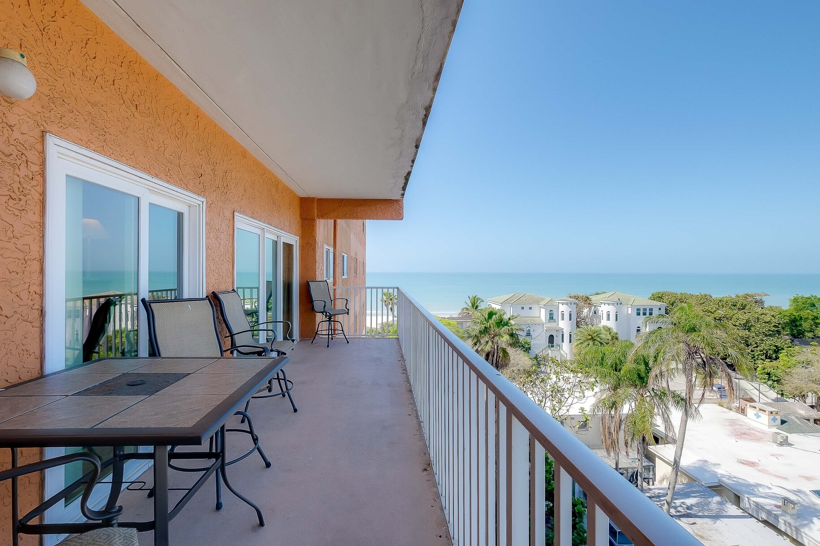 A balcony with a table and chairs, overlooking a coastal area with buildings and palm trees.