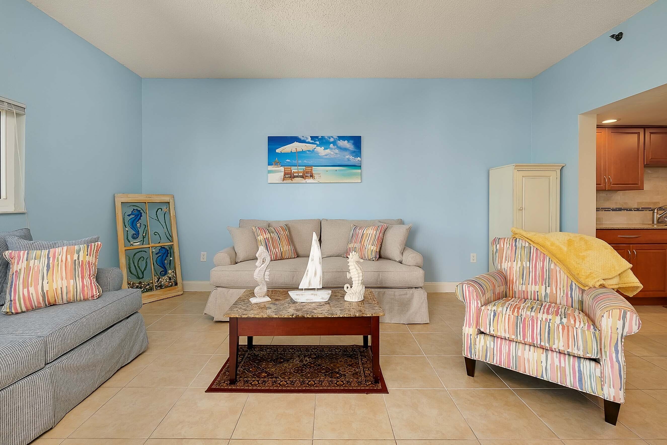 Una sala de estar con sofá, sillón y mesa de centro, un cuadro de una playa y una obra de arte enmarcada en la pared.