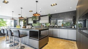 Une cuisine moderne dotée d’un îlot central, de tabourets de bar, de luminaires suspendus et offrant une vue sur la verdure environnante.