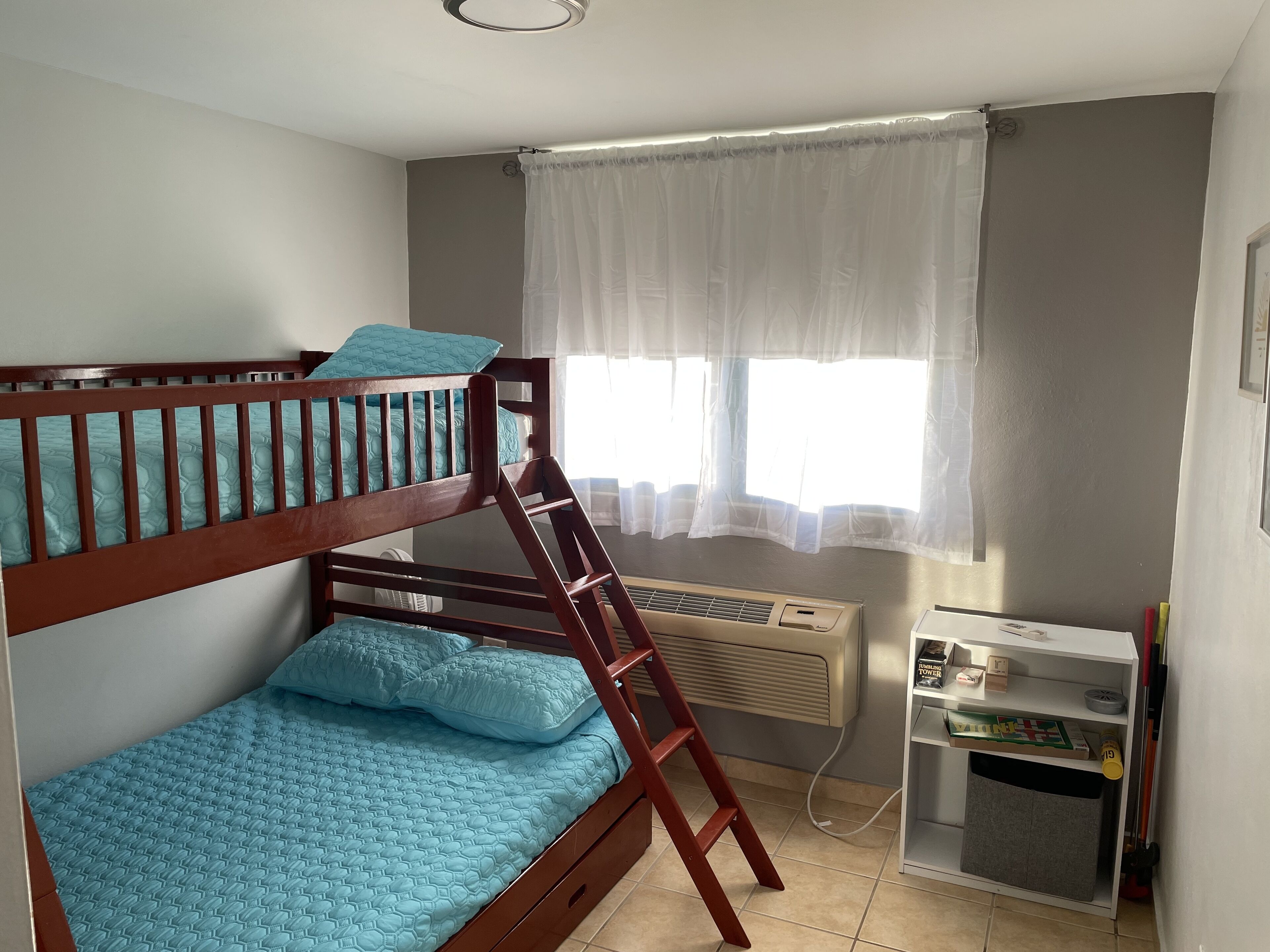 A bunk bed with a ladder, a window with sheer curtains, an air conditioning unit, and a small shelf with books and a toy.