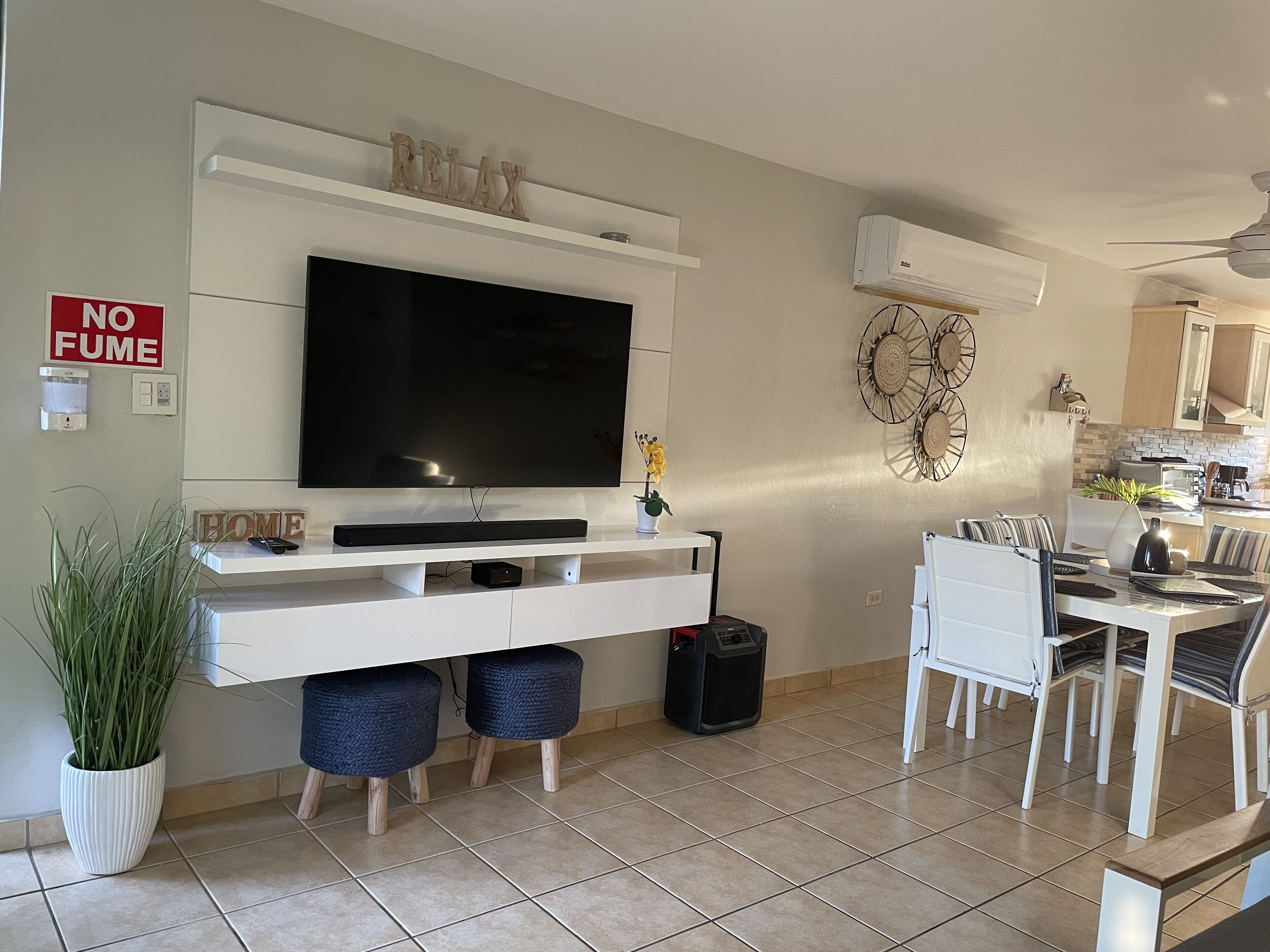 A living room with a flat-screen TV, a dining area with a table and chairs, a white console table with a TV stand, a 'NO FUME' sign, a wall-mounted air conditioner, and a ceiling fan.