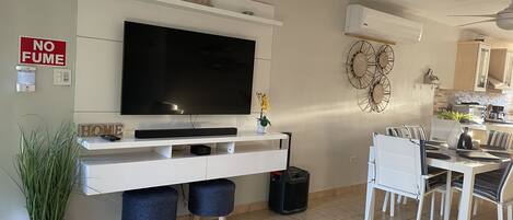 A living room with a flat-screen TV, a dining area with a table and chairs, a white console table with a TV stand, a 'NO FUME' sign, a wall-mounted air conditioner, and a ceiling fan.