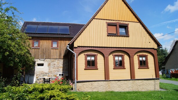 Uma casa de dois andares com fachada amarela e detalhes em marrom, fundação de pedra e telhado inclinado com painéis solares.