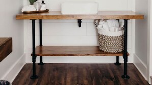 Un baño con pared de azulejos blancos, tocador de madera con lavabo, espejo y una tubería negra.