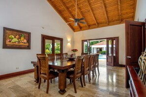 Uma sala de jantar com teto de madeira, uma mesa grande e cadeiras. Há um quadro na parede e uma porta que dá para o exterior.