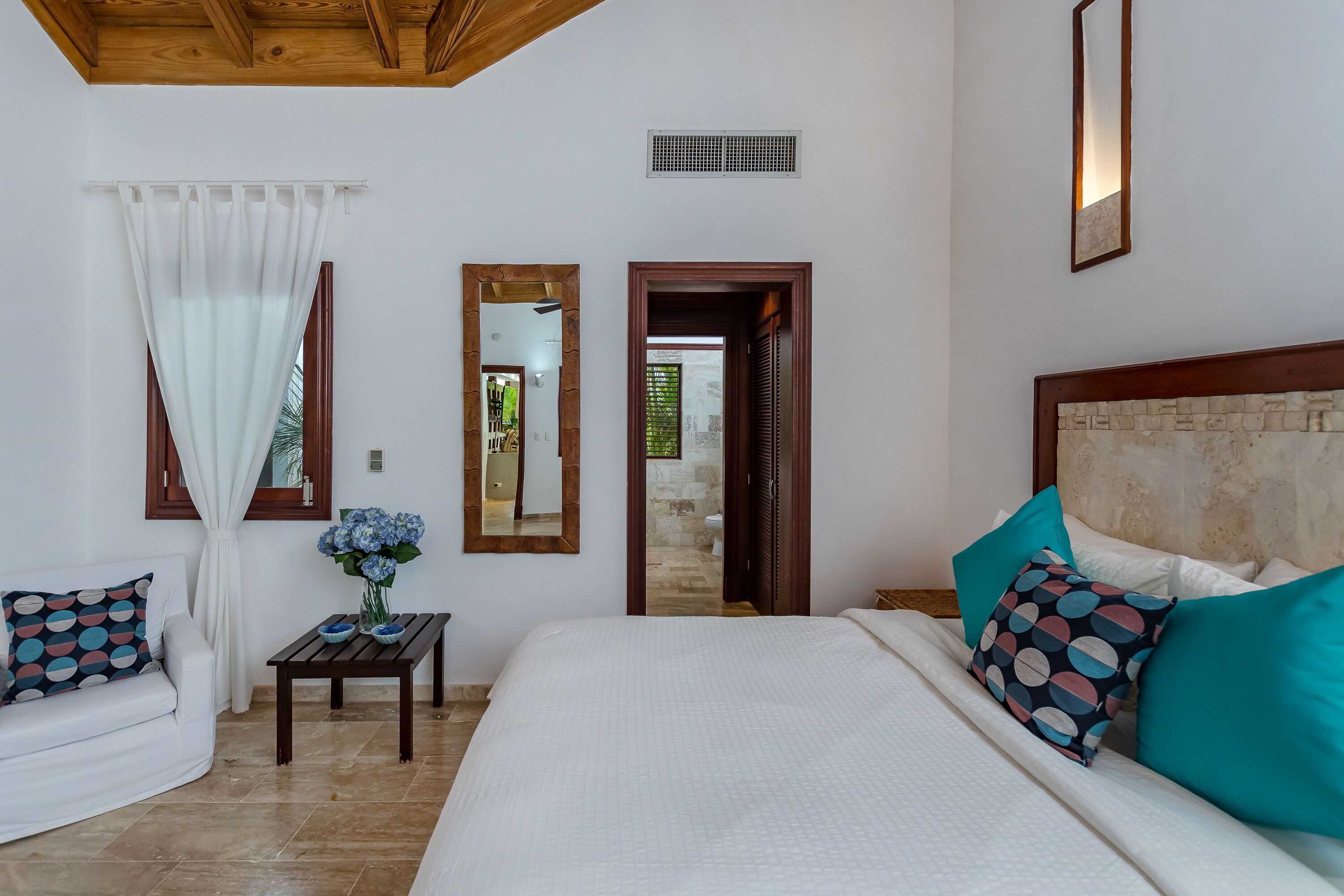A bedroom with a large bed, a wooden headboard, a white sofa, a small table with a vase, and a mirror on the wall.