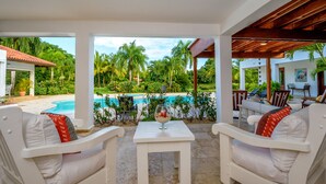 A covered outdoor seating area with white furniture, a glass table with a candle, and a view of a swimming pool and tropical vegetation. - Luxurious Casa de Campo Villa for up to 12 people (La Romana)